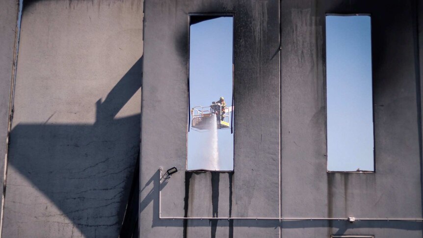 A view through a window of a firefighter spraying water on a fire from a crane.