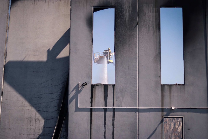 A view through a window of a firefighter spraying water on a fire from a crane.