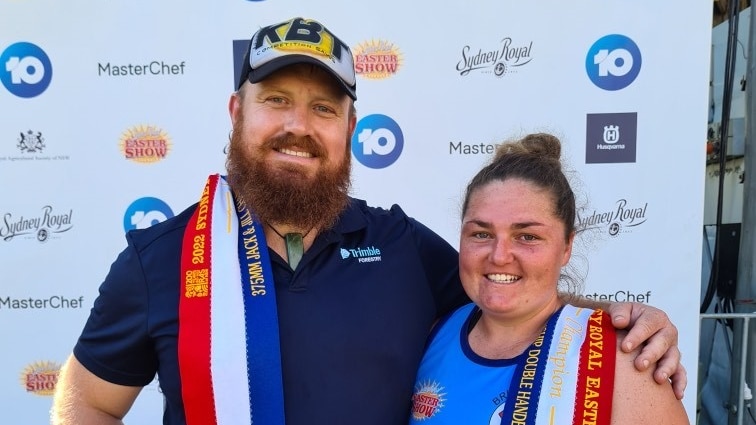 A man and woman stand in front of a banner with award ribbons over their shoulders