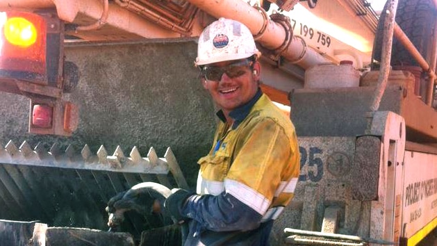 Rhys Connor, dressed in high-vis gear, stands in front of a large piece of mining machinery.