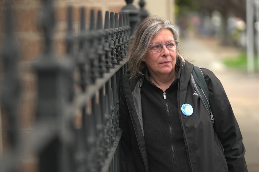 A woman with glasses wearing dark top leaning against a iron fence