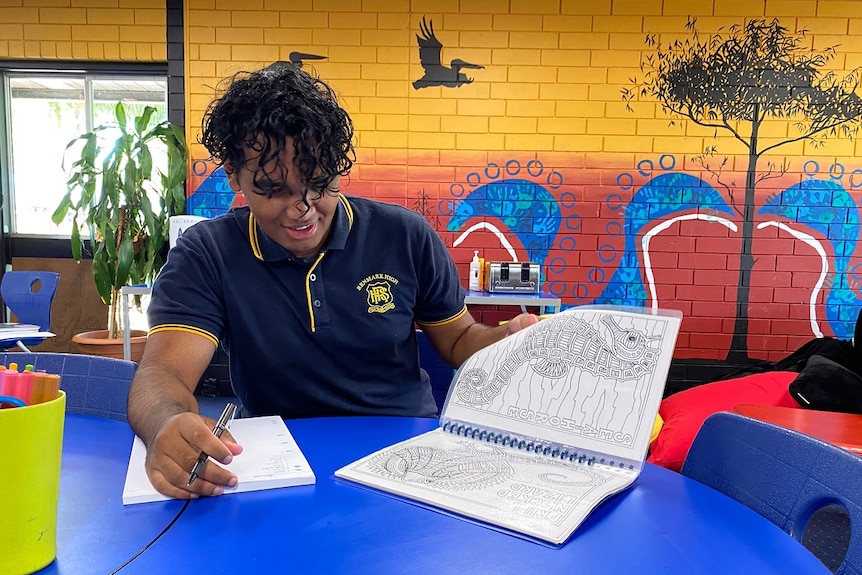 A young man is sitting at a desk looking at an open folder. There is a colourful mural on the wall behind him.