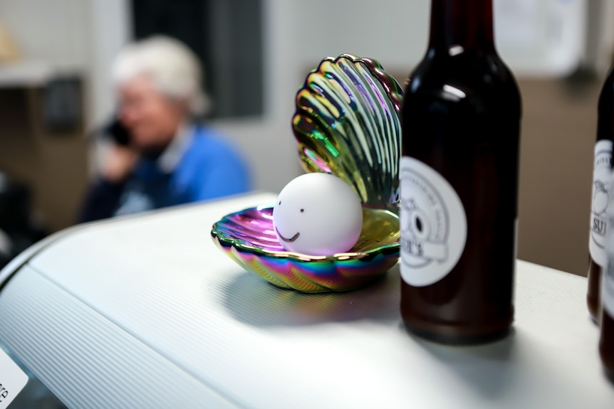 A smiling drawn on face on a fake plastic clam sits atop a shop counter with a woman on the phone in background