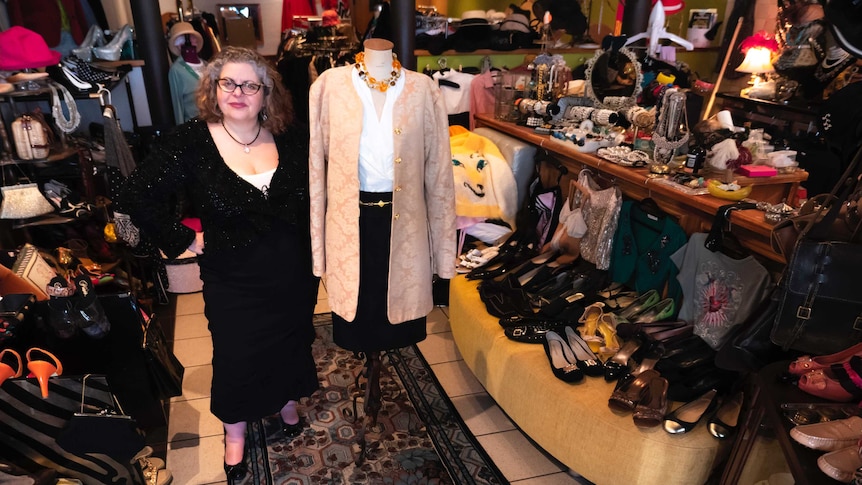 A woman dressed in a black outfit stands next to a fashionably-dressed mannequin in a busy vintage shop.
