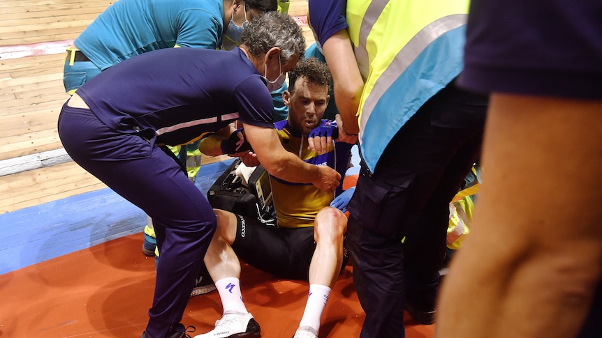 An injured elite cyclist sits in distress on the side of a track as medical staff try to help him to his feet after a crash.