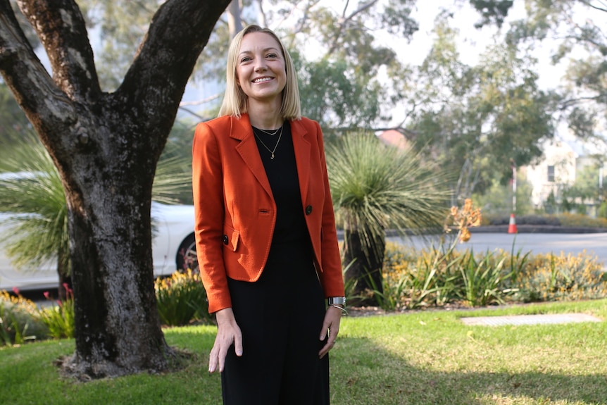 A wide shot showing Mia Davies standing in the Fern Garden outside Parliament House wearing a red jacket and black pants.