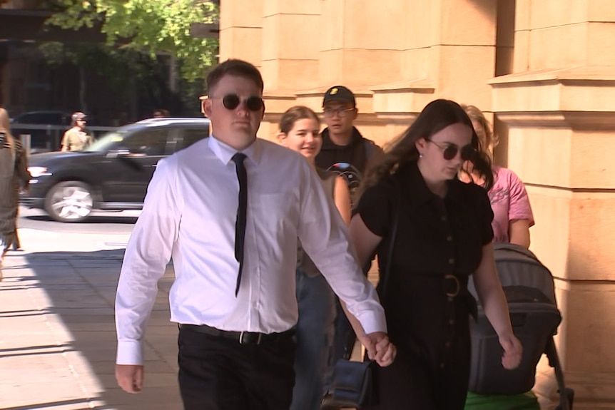 Cameron Brodie-Hall looks at the camera as he walks to court while holding a woman's hand.
