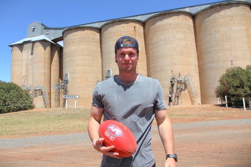 A man holds an AFL football in front of large silos.