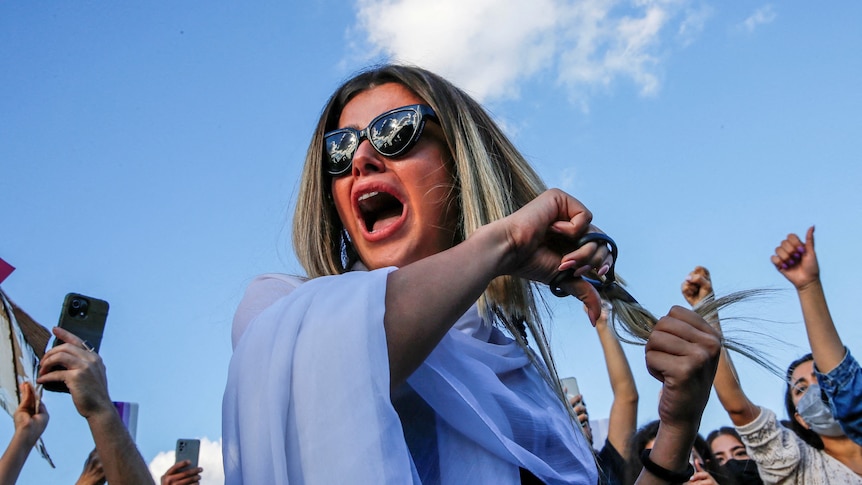 A woman holds her long hair and scissors to it outside in a protest.