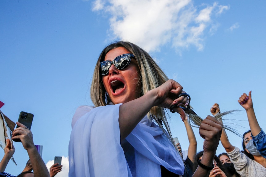 A woman holds her long hair and scissors to it outside in a protest.
