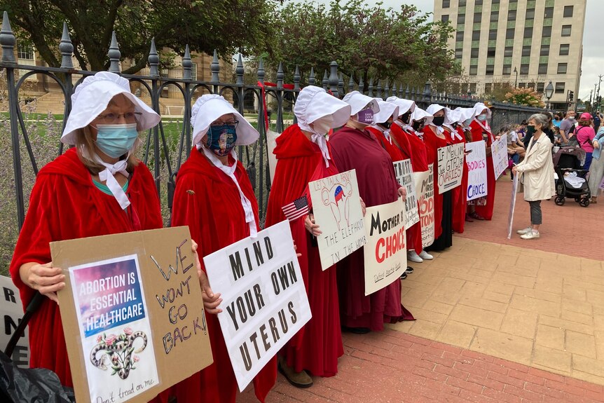 women dressed in red and white outfits of characters from the Handsmaid's Tale series stand in a line with placards 