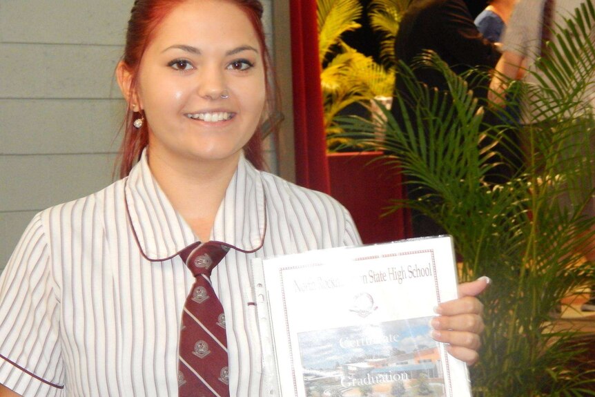 A teenage girl with brown hair and eyes smiles as she holds a graduation certificate