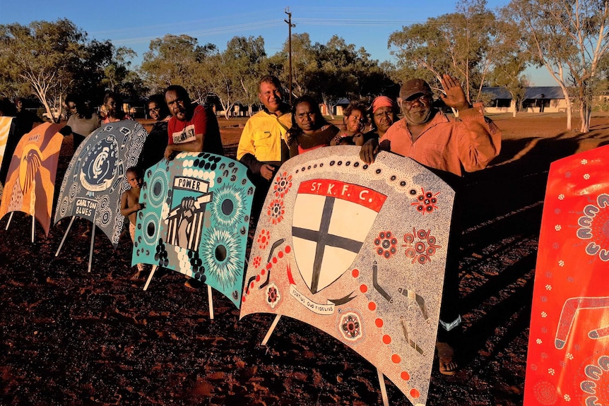 A group of Indigenous residents standing with several bonnets painted with AFL team logos.