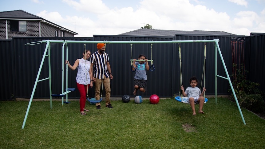 A woman and man watch as their two young children play on a swing set in a backyard.