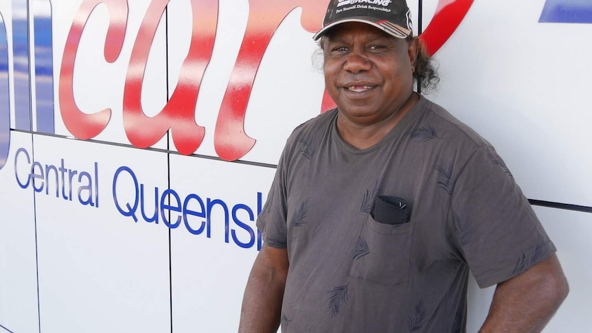 An Aboriginal man wearing a cap and a big smile stands in front of a sign that says Anglicare Central Queensland.