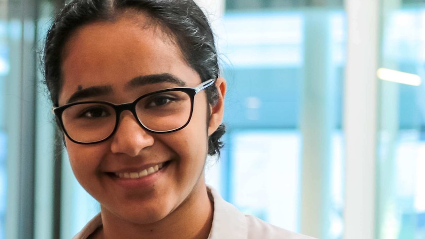 A teenage girl wearing glasses and a white shirt looks at the camera, smiling.