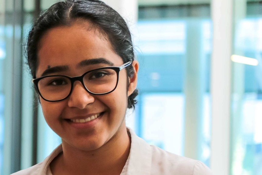 A teenage girl wearing glasses and a white shirt looks at the camera, smiling.