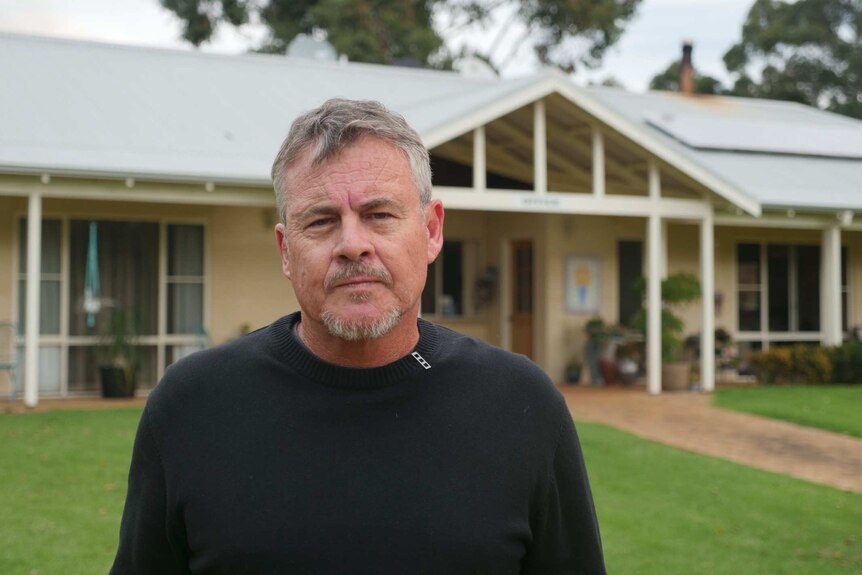 A disappointed-looking man in a black jumper stands in front of the entrance to cottage accomodation.