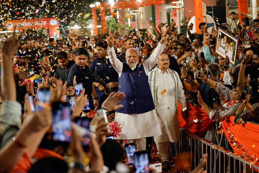 Narendra Modi walks through confetti surrounded by hundreds of supporters holding his hands in the air making a peace symbol