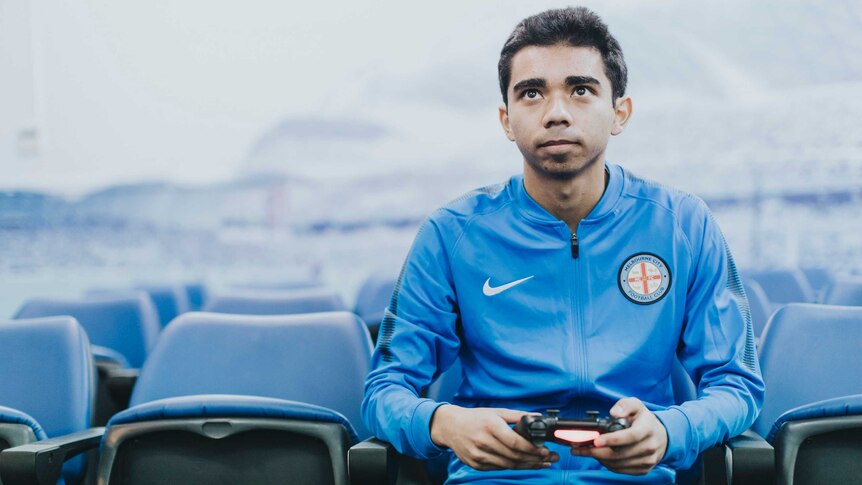 Marcus Gomes sitting in the stands wearing a blue Melbourne City jumper and holding a PlayStation controller.