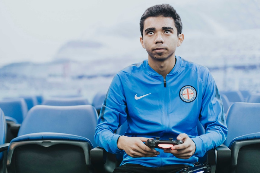 Marcus Gomes sitting in the stands wearing a blue Melbourne City jumper and holding a PlayStation controller.