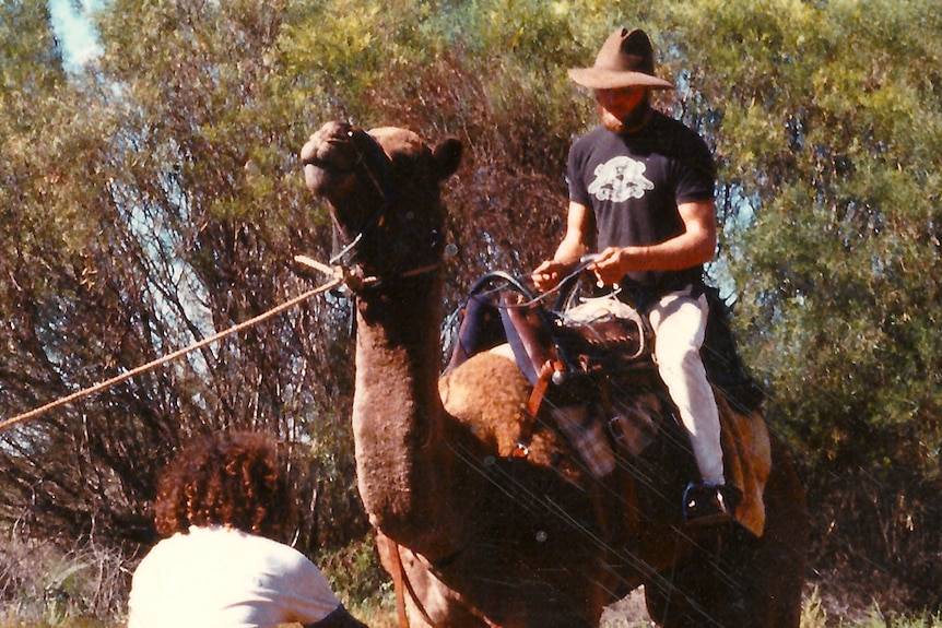 A bearded man sits on a camel as another man stands with a lasso ready to capture the camel.