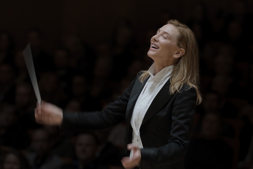 A brown-haired, 50-something woman smiles broadly, her eyes closed, as she moves a baton through the air