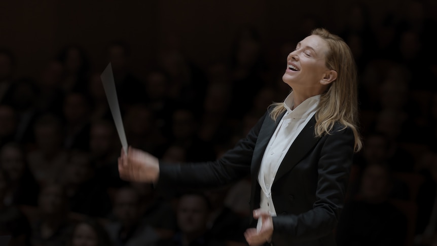 A brown-haired, 50-something woman smiles broadly, her eyes closed, as she moves a baton through the air