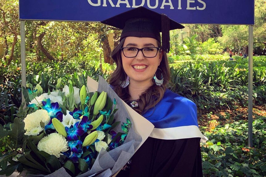 Drisana is wearing a mortarboard and university robes and holding a large bunch of flowers at her uni graduation.