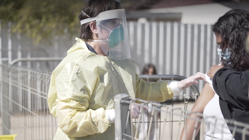 A nurse in PPE administers a vaccine to a woman over a fence.