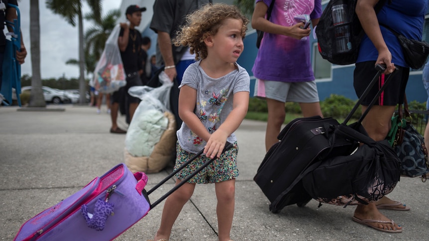 A three-year-old girl wheels a pink and purple suitcase while following her parents into a shelter.