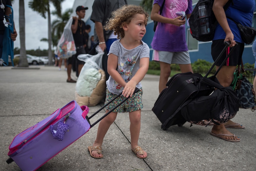 A three-year-old girl wheels a pink and purple suitcase while following her parents into a shelter.