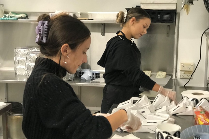 Two women wearing black pack cheese in white paper.