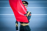A young man in Chinese military dress stands to attention with a red flag blowing across his face