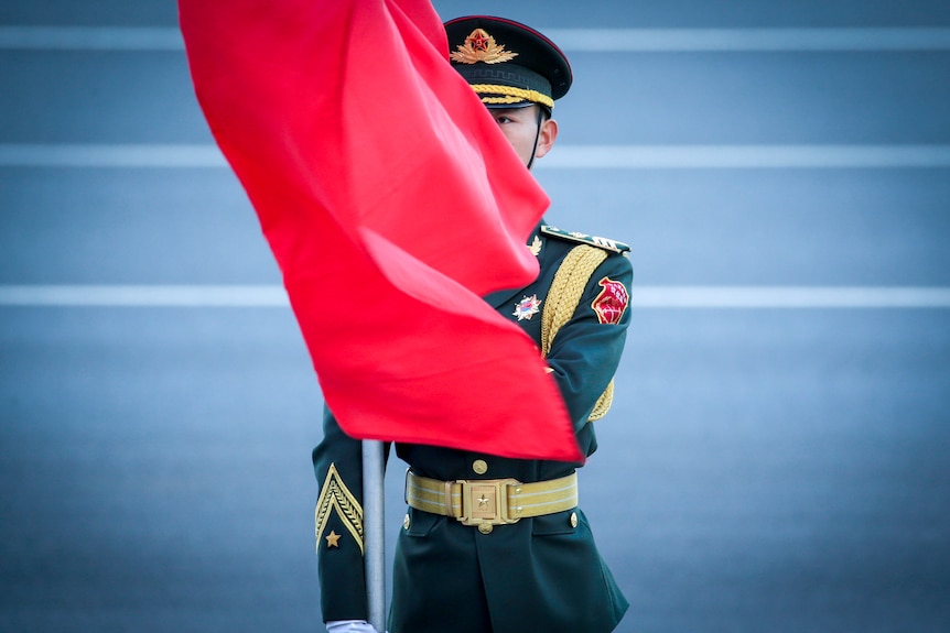 A young man in Chinese military dress stands to attention with a red flag blowing across his face