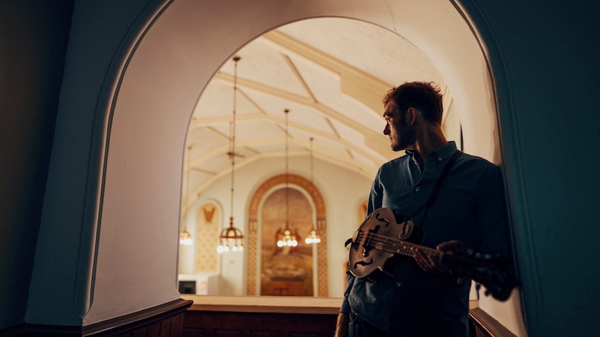 Chris stands in the doorway of a church, he is looking back and holding a mandolin.