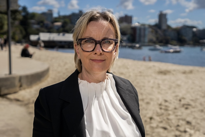 Michele Carnegie wears thick rimmed glasses and smiles at the camera at the beach.