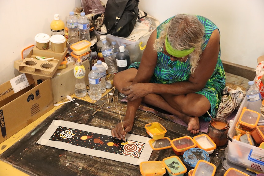 A woman sitting on the floor of a room and painting a canvas in a traditional Aboriginal style. 