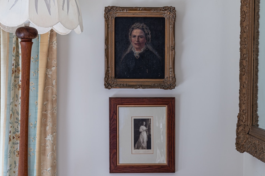 One portrait and one photo of women hang above each other on a white wall. 