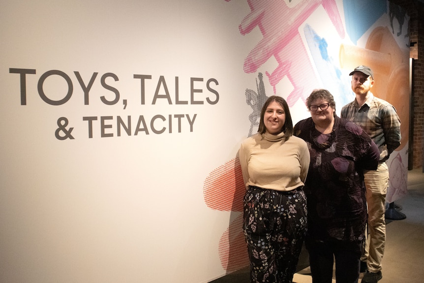Three people stand in front of a wall with Toys, Tales and Tenacity title written on it