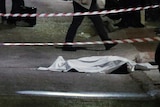 A cadaver is covered by a sheet and guarded by police on the side of a dark road near a construction skip bin