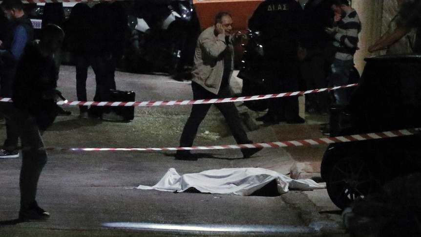A cadaver is covered by a sheet and guarded by police on the side of a dark road near a construction skip bin