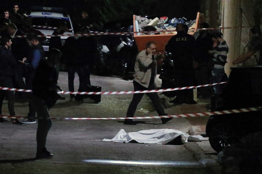 A cadaver is covered by a sheet and guarded by police on the side of a dark road near a construction skip bin