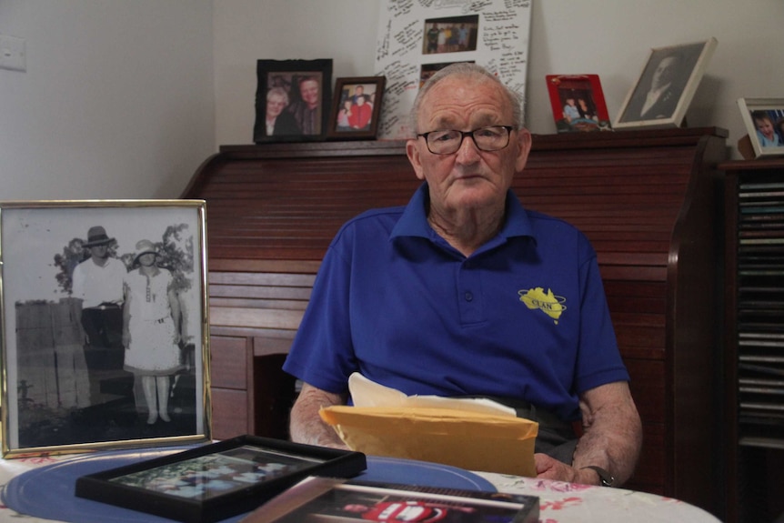 An older man with glasses and a blue shirt, sitting at a table with photos and documents.