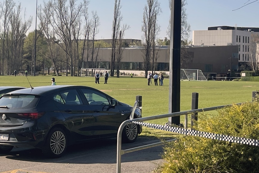 Cars parked next to a football pitch. Police tape is stretched across part of the area. Several police are on the pitch.