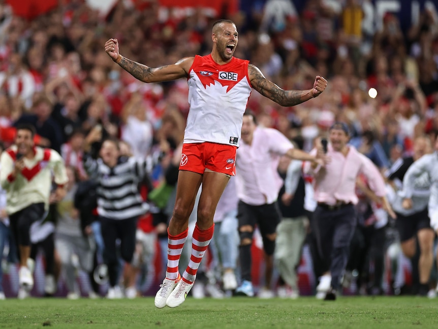 AFL superstar Lance Franklin jumps in the air with arms spread in triumph, as many fans sprint towards him on the ground.