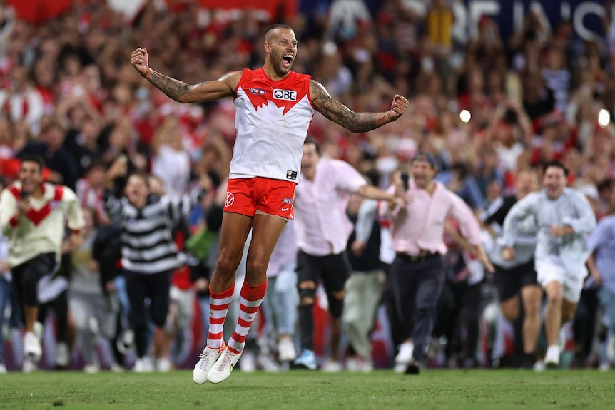 AFL superstar Lance Franklin jumps in the air with arms spread in triumph, as many fans sprint towards him on the ground.