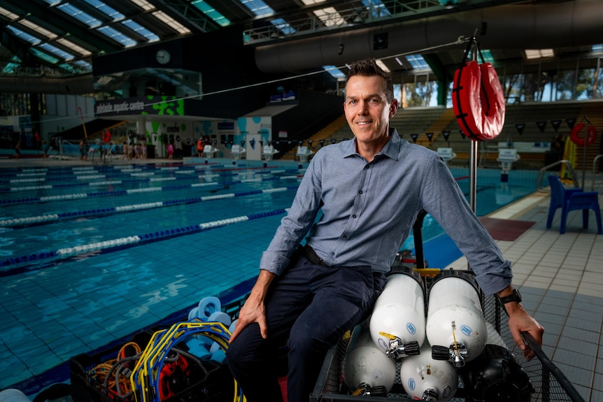 A man with short brown hair wearing a blue shirt sitting next to a swimming pool inside an aquatic centre.