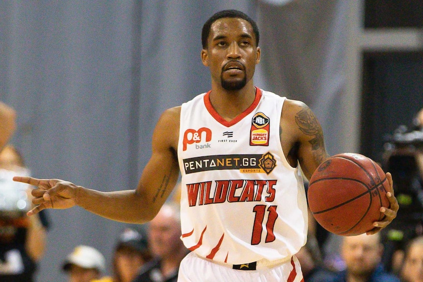 Bryce Cotton dribbles the ball up court in his white Perth Wildcats uniform with his right arm outstretched making a sign.