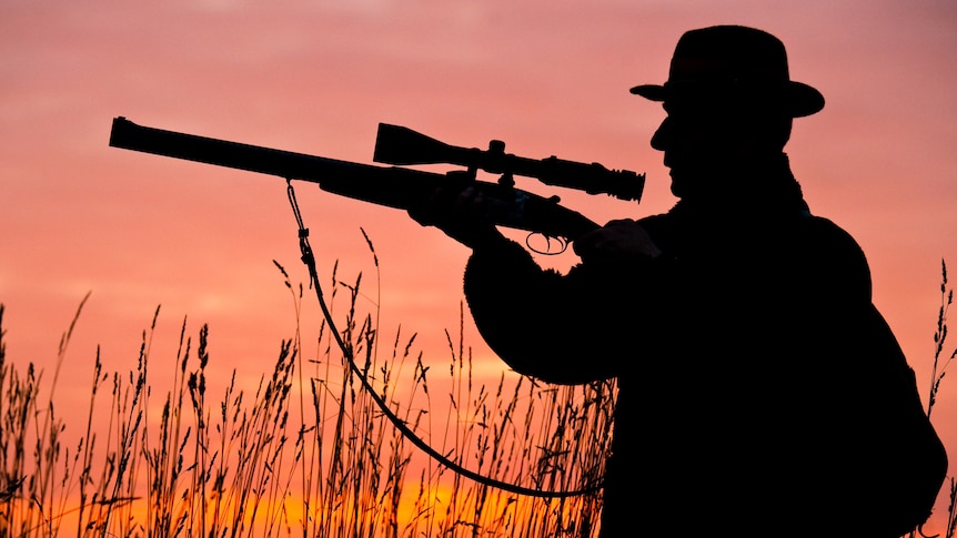 Silhouetts of a hunter aiming his rifle through a telescopic viewfinder as the sun sets behind him.